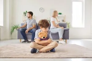 Frustrated upset boy son sitting on floor with toy bear while his mother and father sitting on opposite sides of sofa ignoring each other after quarrel at home. 