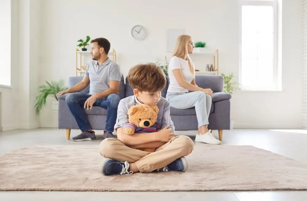 Frustrated upset boy son sitting on floor with toy bear while his mother and father sitting on opposite sides of sofa ignoring each other after quarrel at home.